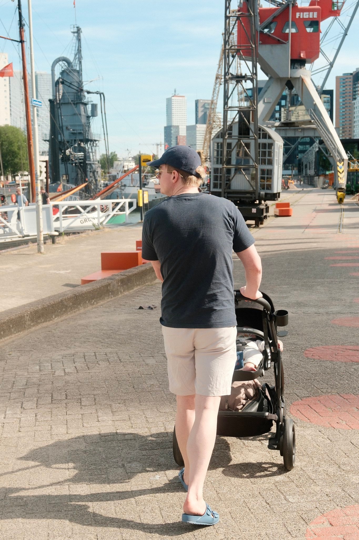 gallery-01 Man in a navy cap walking with a baby stroller along a harbor promenade with cranes in the background.