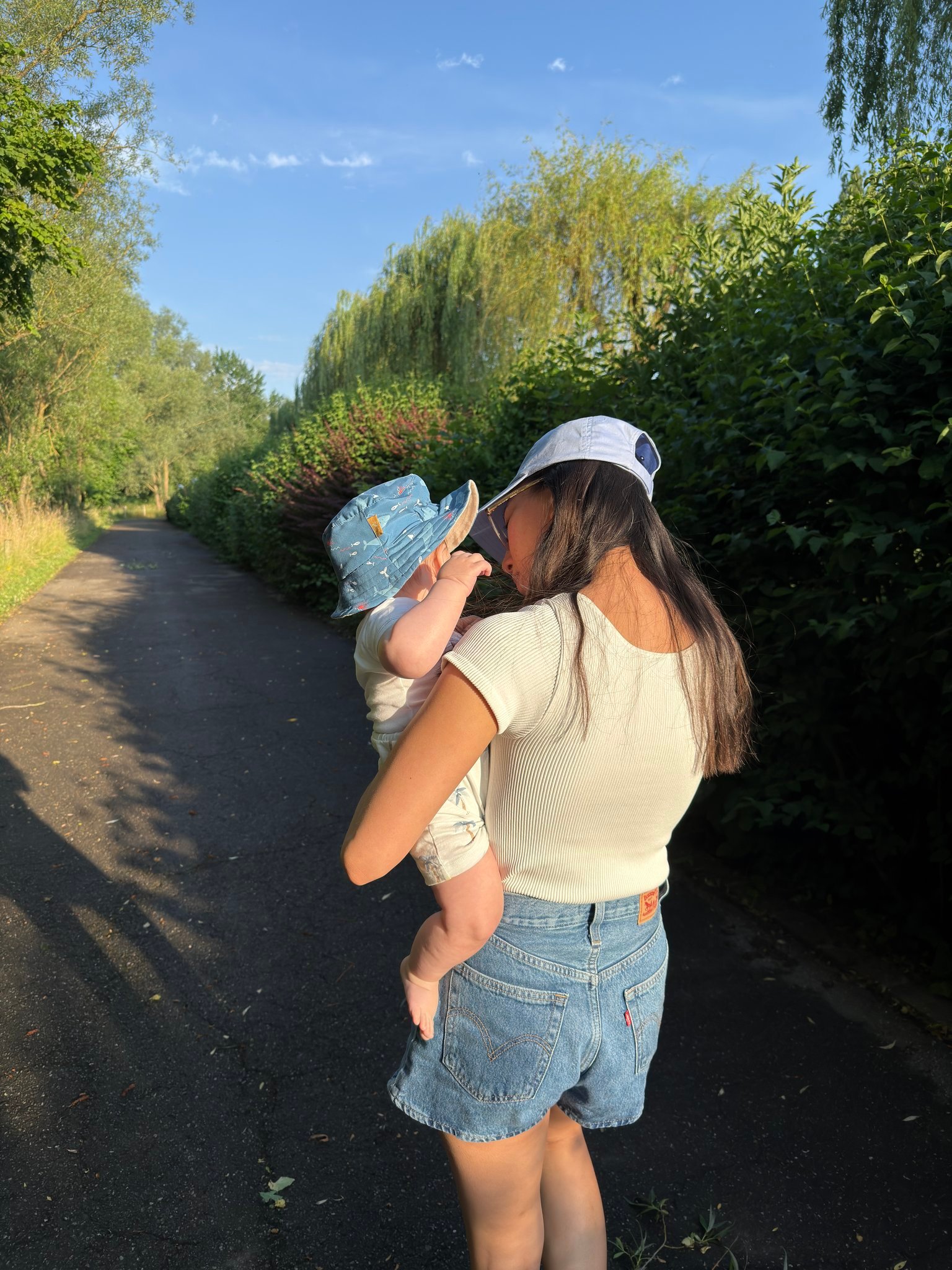 Woman carrying a small child along a shaded park path with green hedges and trees on a sunny day, front-right view from behind a mother.