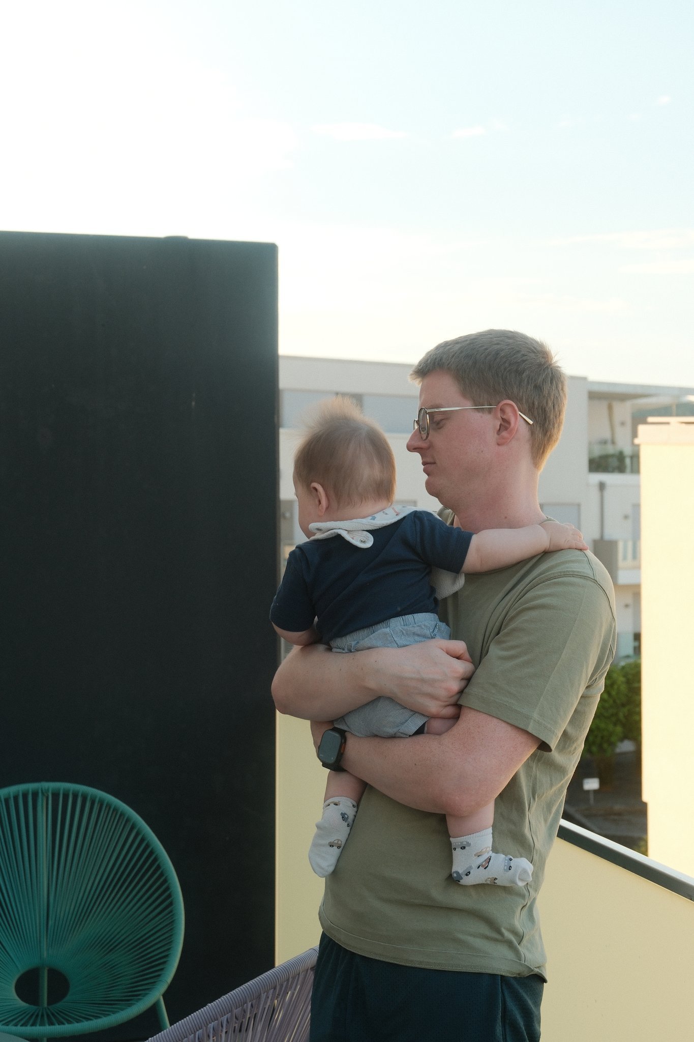 Man wearing glasses holds a baby on a balcony, facing right, with a teal chair nearby and modern buildings in the background.