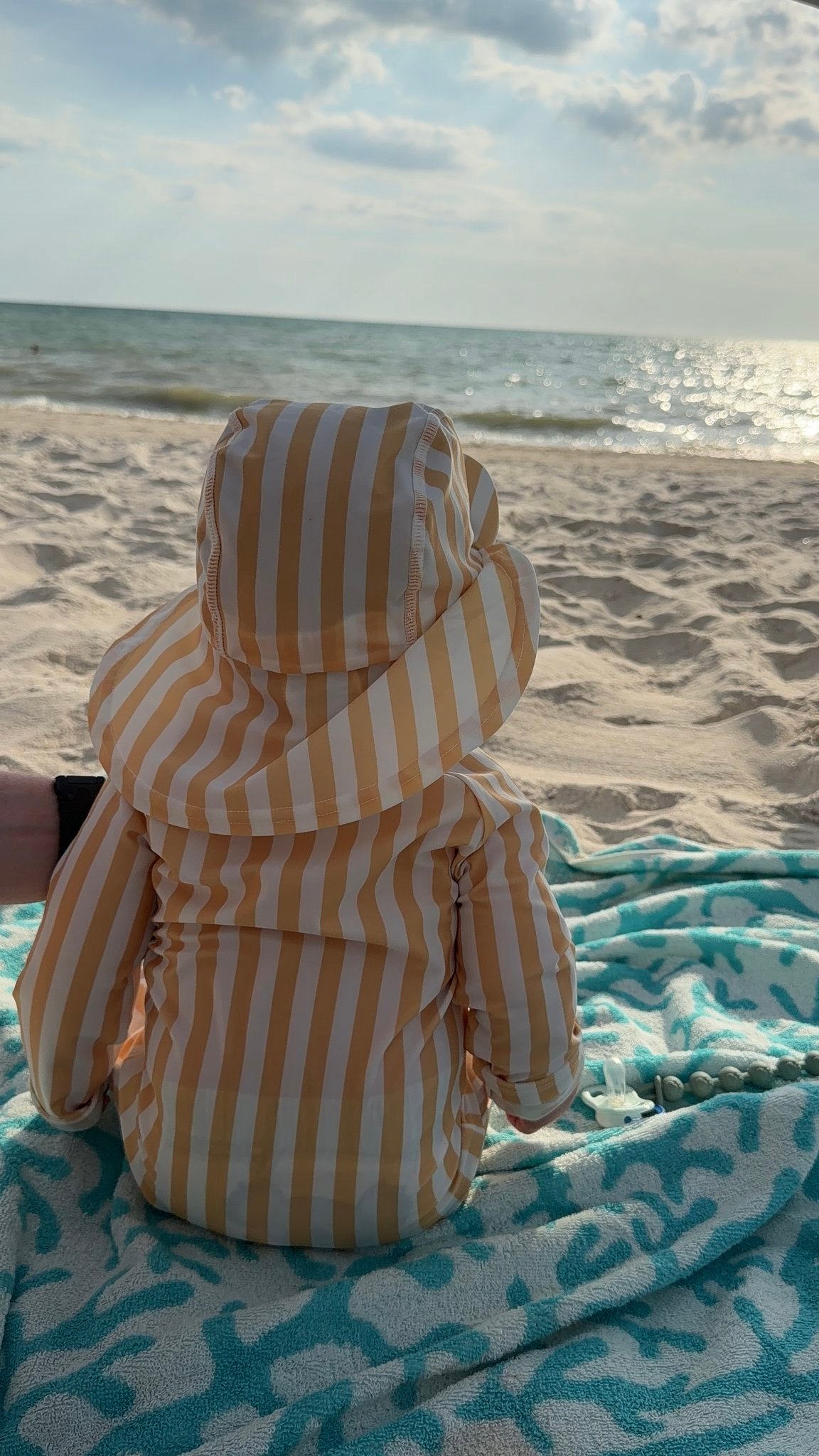 Child in beige-and-white striped sun hat sits on a blue patterned towel on the sandy beach, gazing at the ocean.