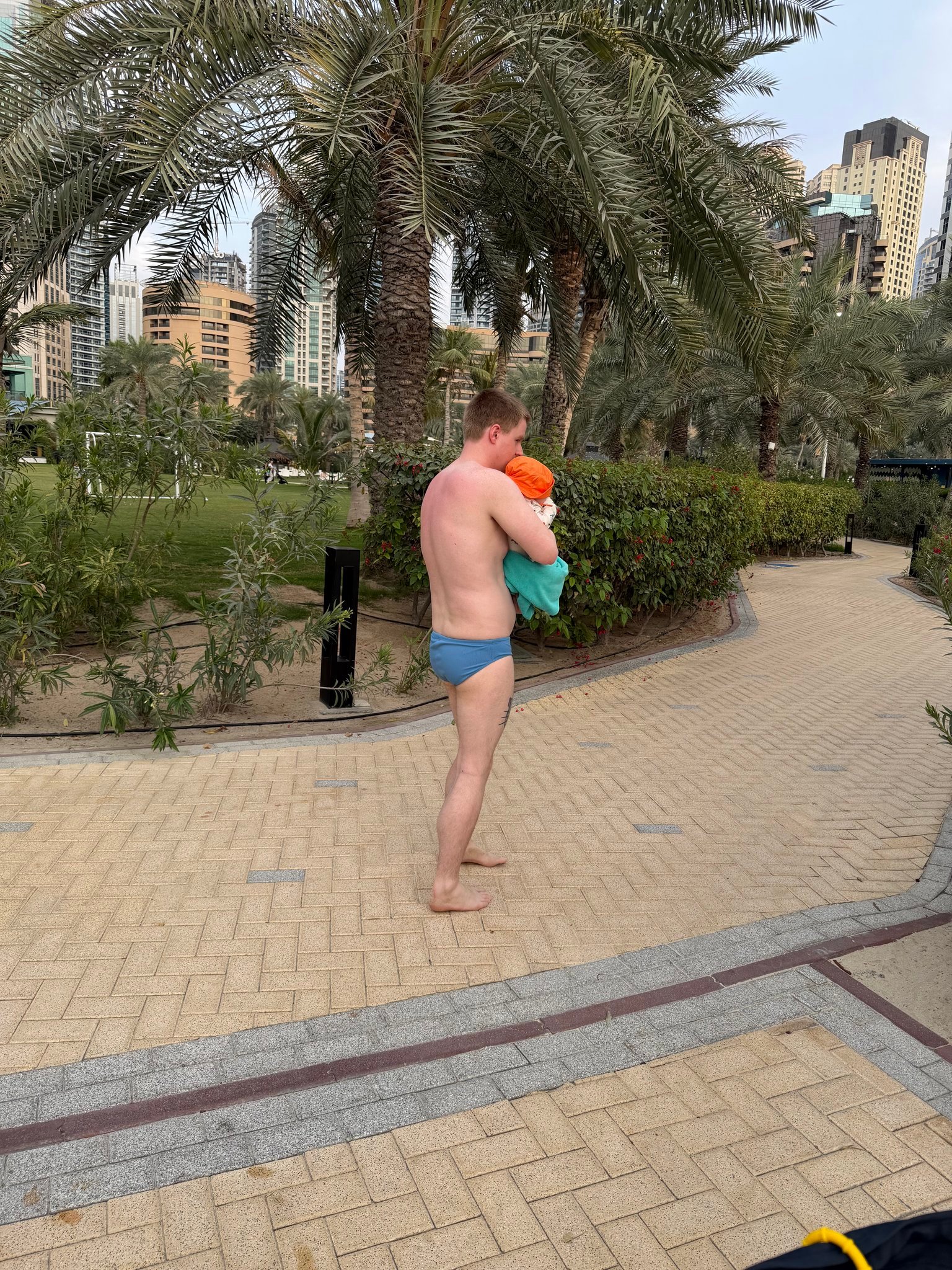 gallery-02 A shirtless man in blue swim trunks holding a baby wearing an orange hat outdoors among palm trees and city buildings in the background.